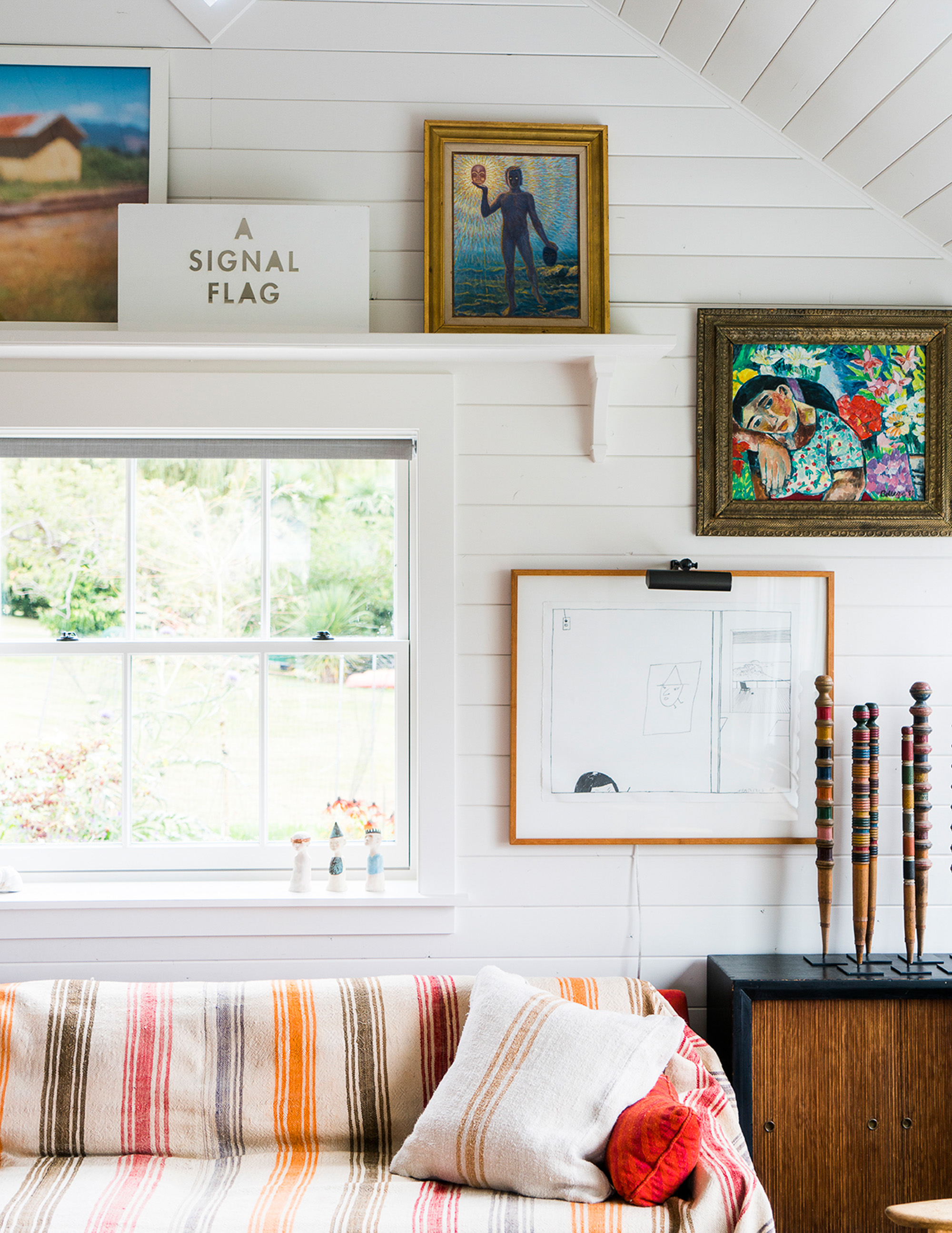 The living room vignette features a cozy sofa covered with a beach blanket, an antique side table with croquet pins, and shiplap white walls adorned with various framed artworks, creating a coastal-inspired and eclectic ambiance.