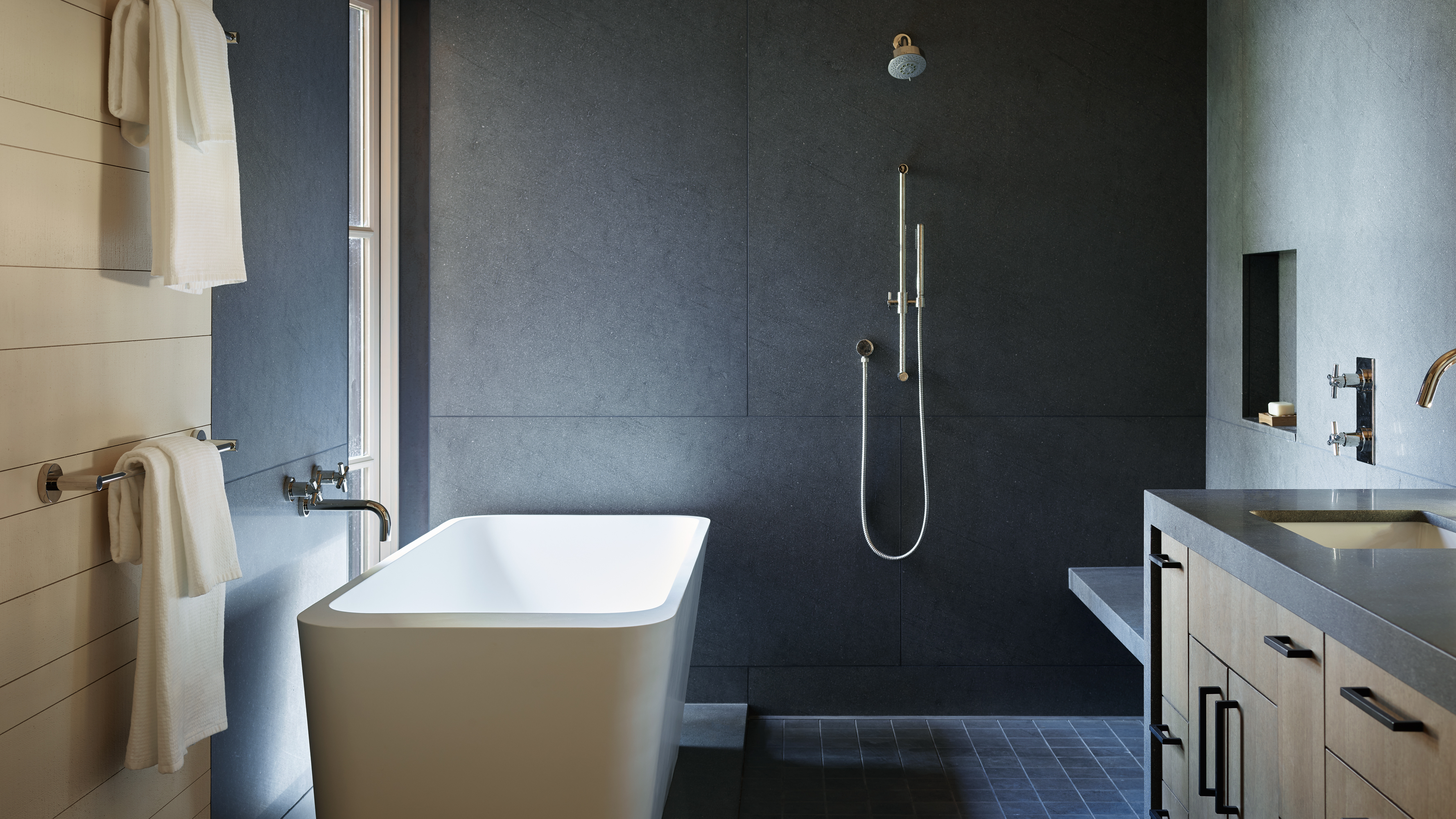 The deep gray, minimal, and sleek bathroom occupies the stone bar at the rear of the house. It projects beyond to offer a view of the water from the bath and shower.