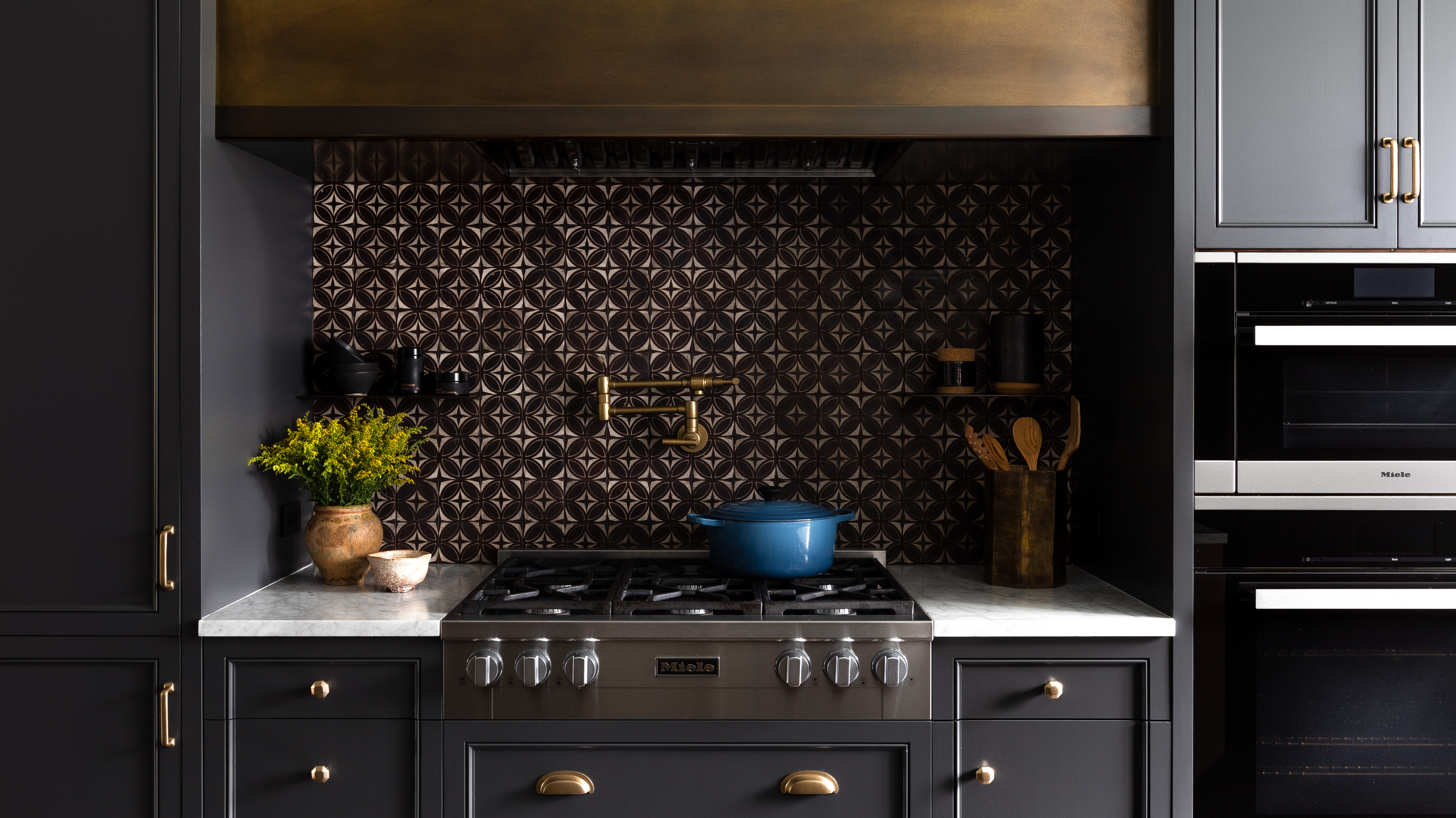 The range area showcases Miele appliances, graphic black and white tile, a custom aged brass hood, dark gray cabinets, and a clean white marble countertop, creating a handsome and masculine aesthetic