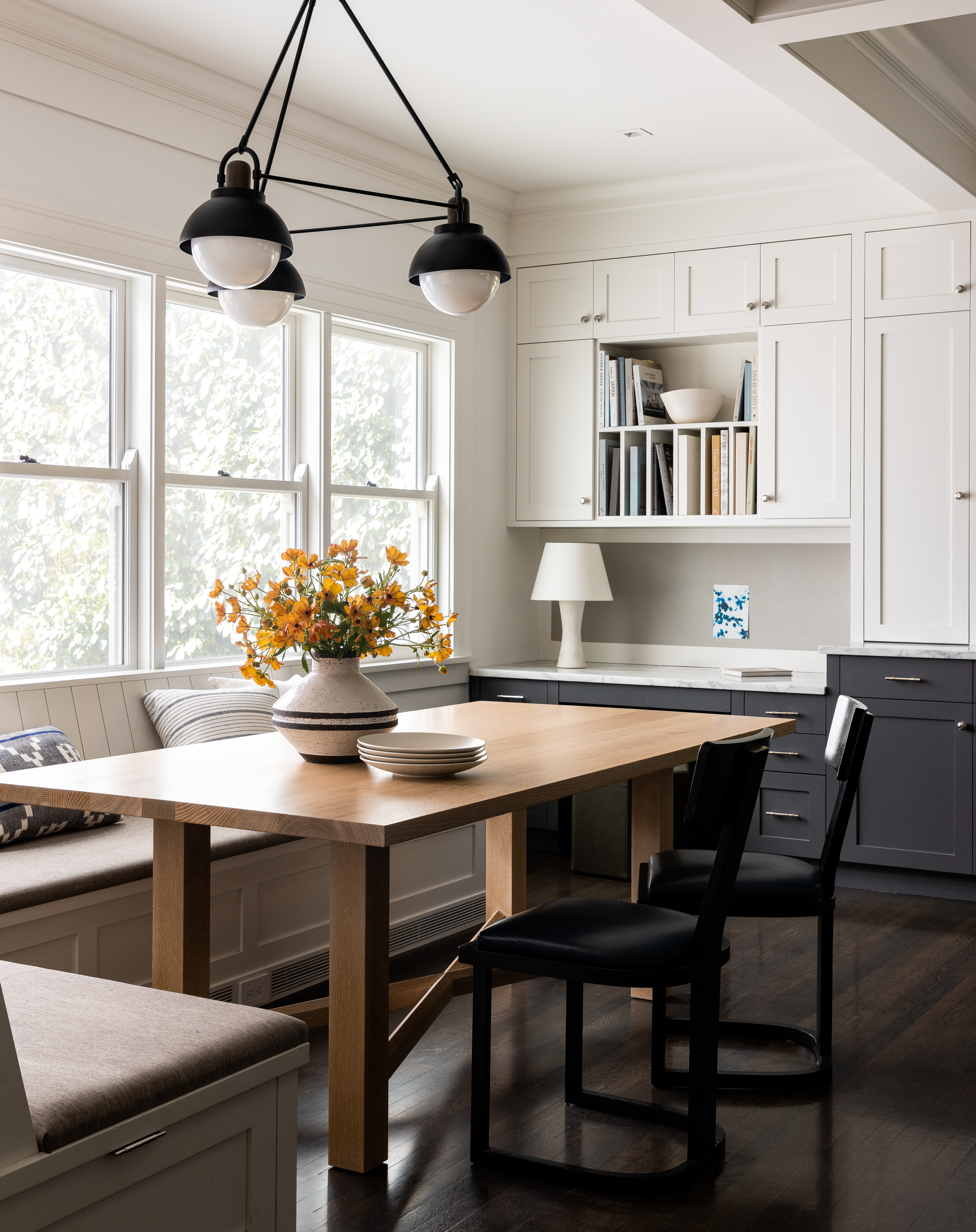 A cozy kitchen nook featuring built-in banquette seating, an Allied Maker light fixture, an oak-finish table, and black-and-white cabinetry.