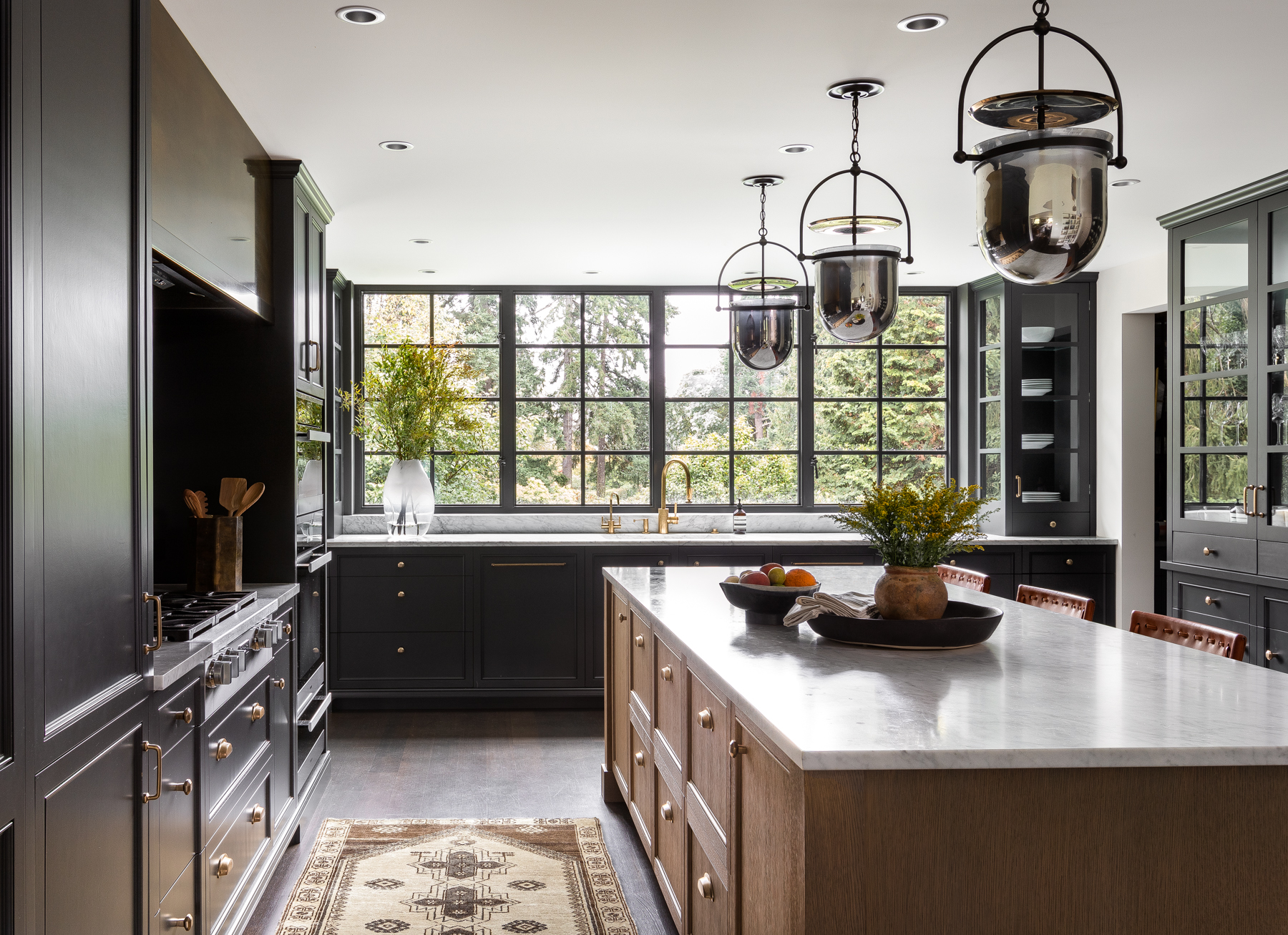 Kitchen featuring a wall of windows above the sink, cool dark gray cabinets, accented oak cabinets, and brass hardware.