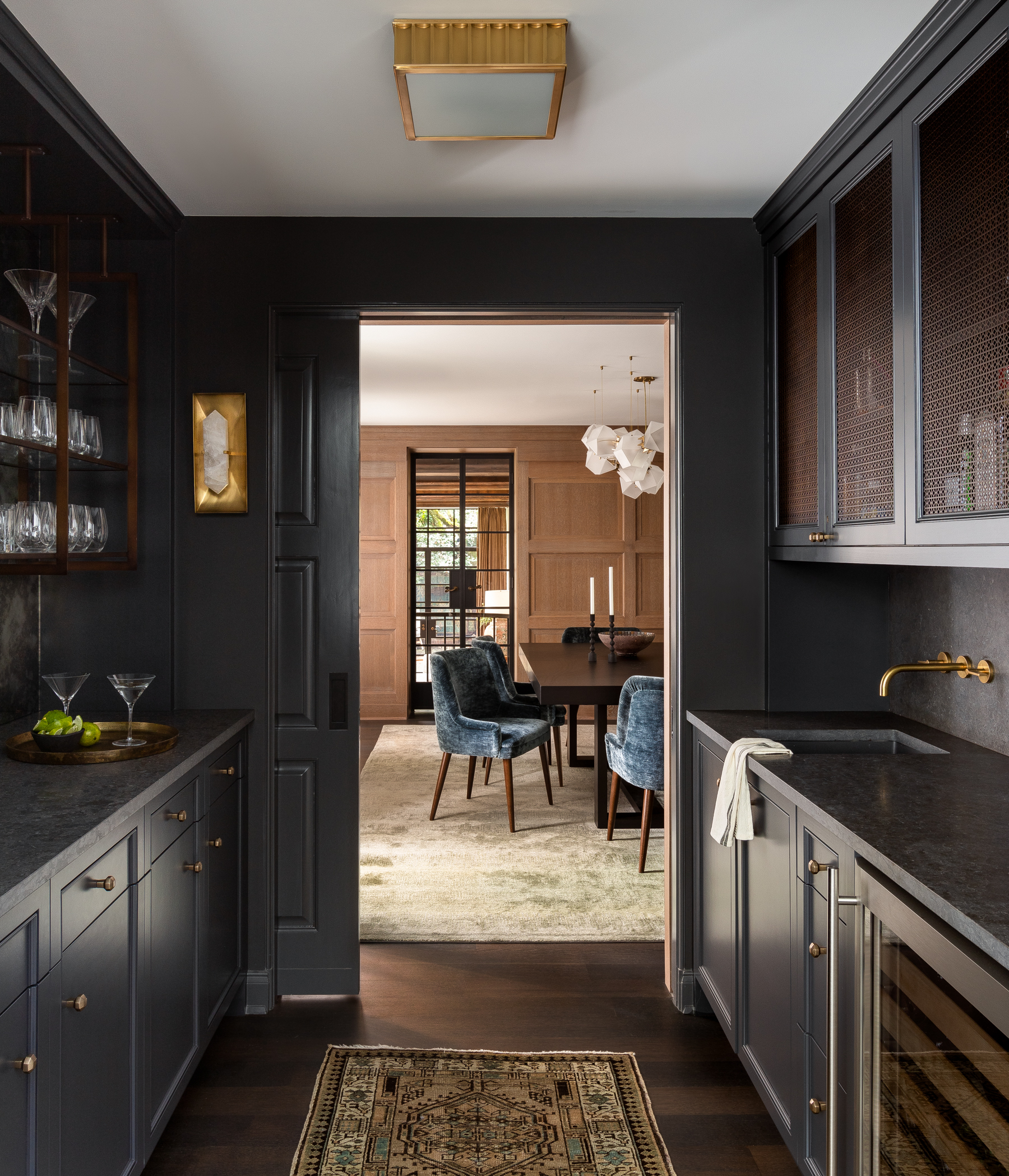Butler's pantry with cool dark gray paint, dark textured marble countertops, brass hardware and fixtures, statement lighting, and an antique mirror backsplash.