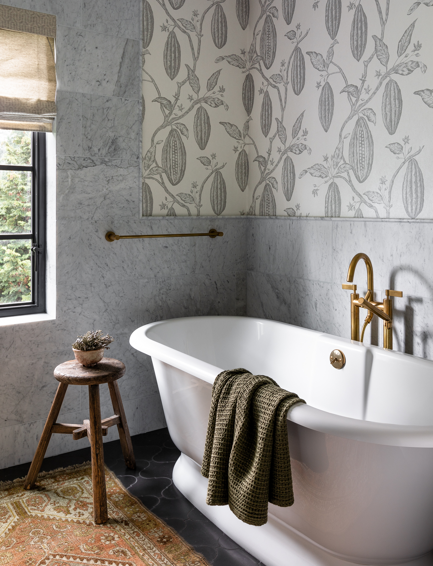 The bathtub lies in a quiet corner of this tranquil space, offering views out the window for a relaxing soak. The intricate botanical wall covering above the marble wainscoting adds to the serene ambiance.