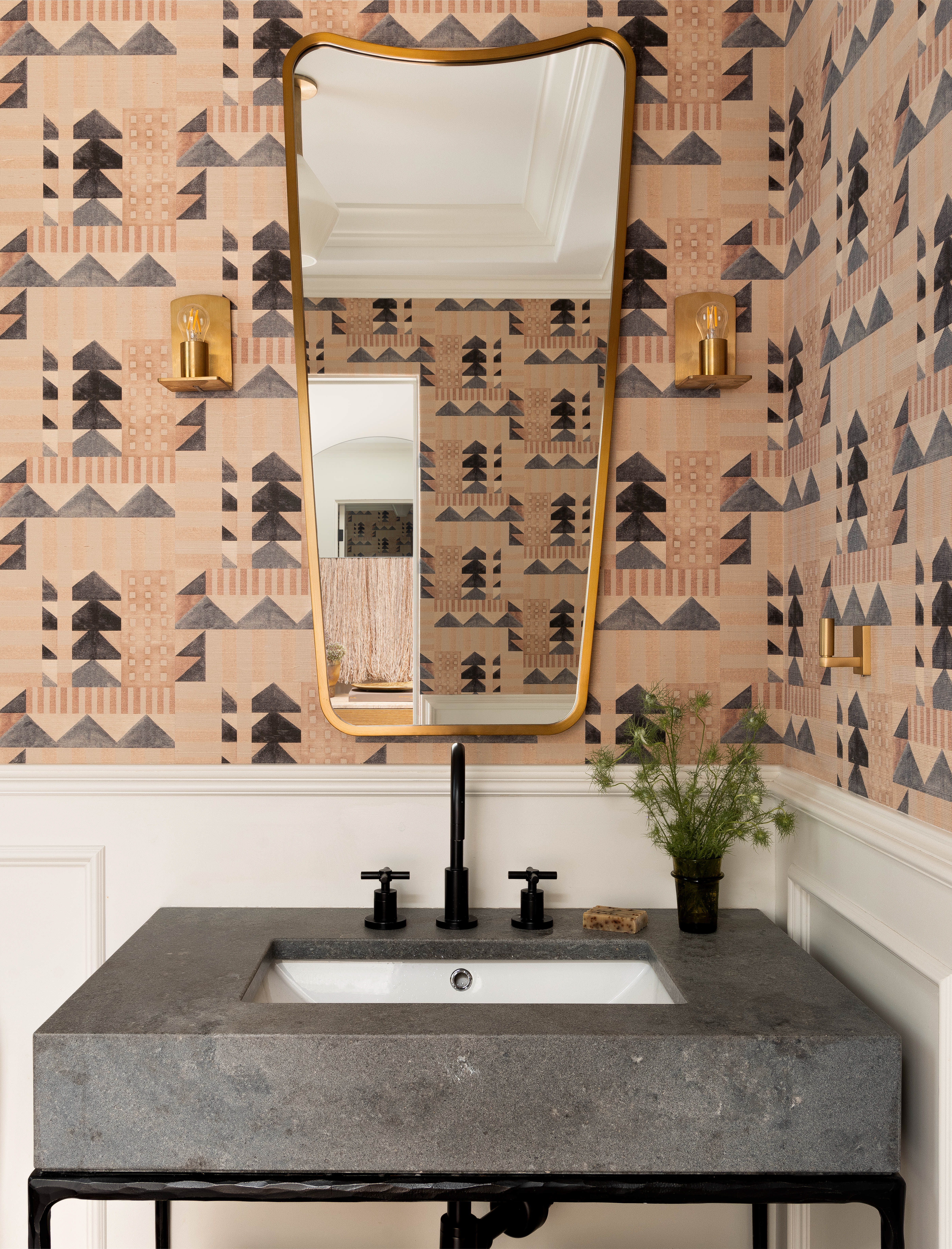 Powder room vanity with geometric wallpaper in dark blues and peach tones. Brass mirror and sconces complement the deep gray marble sink surround.