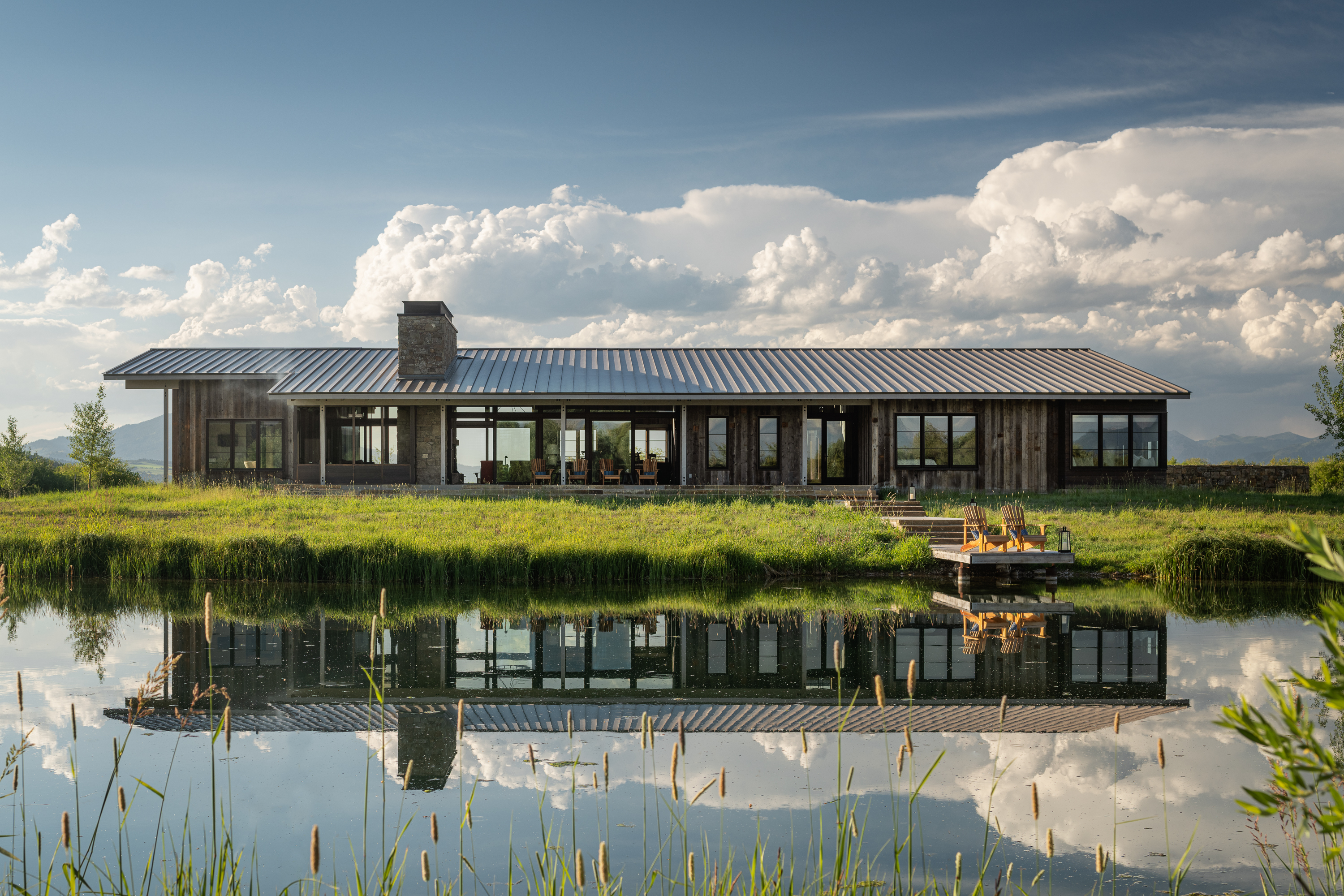 Straight-on shot of Wyoming Retreat featuring reclaimed siding, large windows, covered porch, pathway to the private lake, lush green grass, and dramatic western clouds.