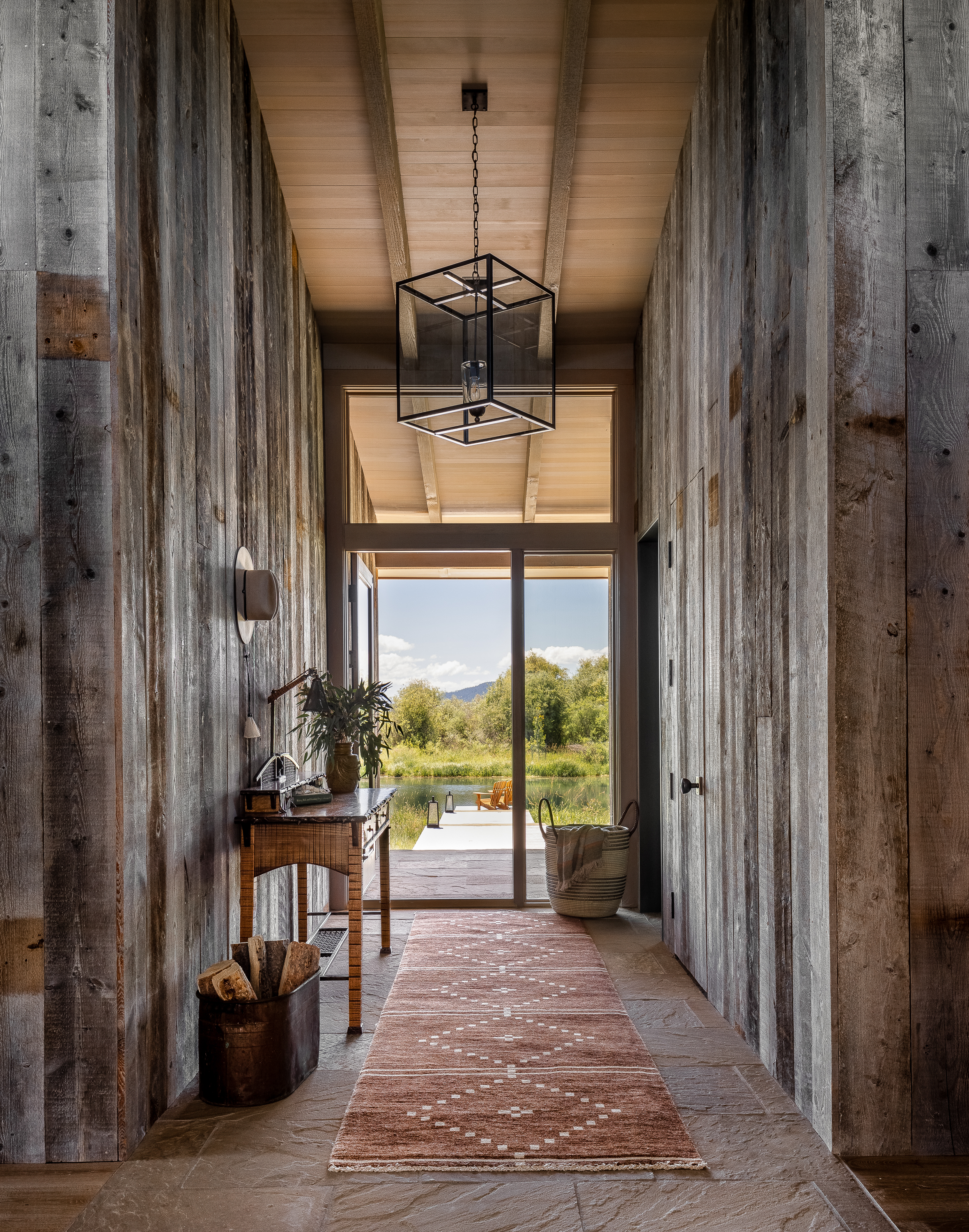 Rustic home interior with reclaimed wood siding, vintage furniture and rugs, and a large-scale metal cube chandelier. The axial hallway leads straight to the lake, offering picturesque views.