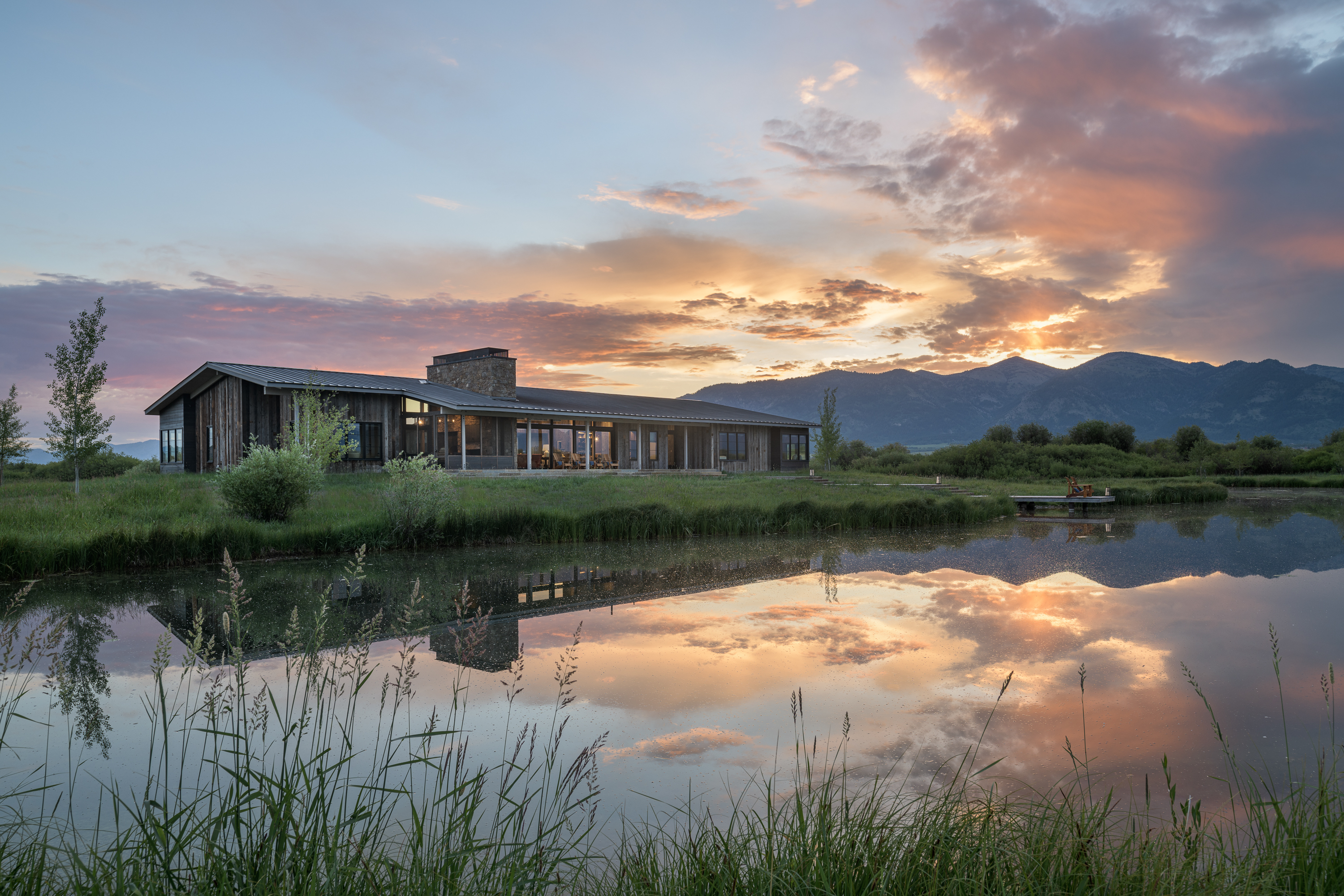Sunset over the Wyoming Retreat, casting a dramatic reflection in the lake. The single-story ranch-style house blends into the serene landscape