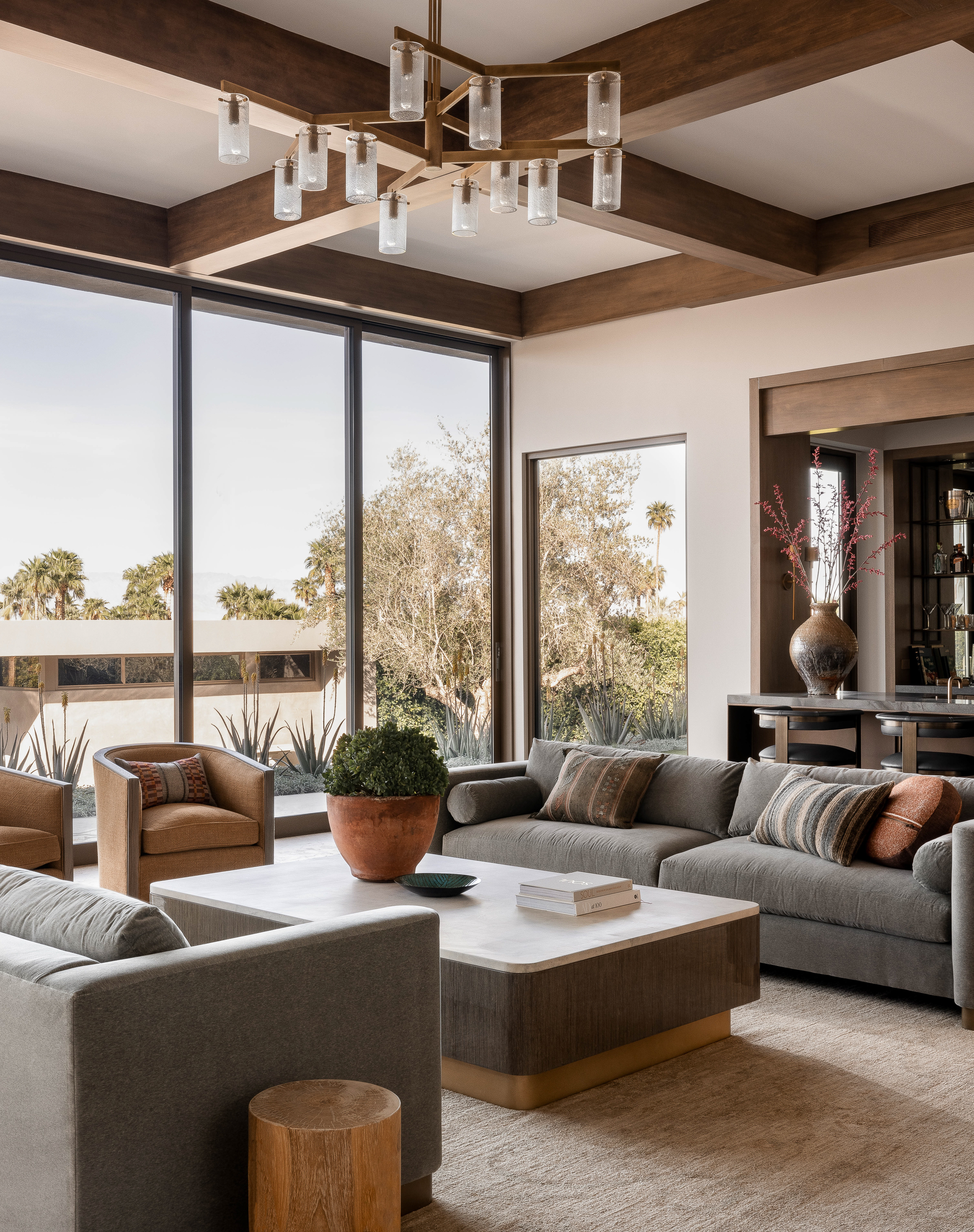 Desert Retreat's living room boasts a bronze fireplace, 10’ velvet sofas, swivel chairs, and floor-to-ceiling glass doors merging indoor-outdoor spaces.