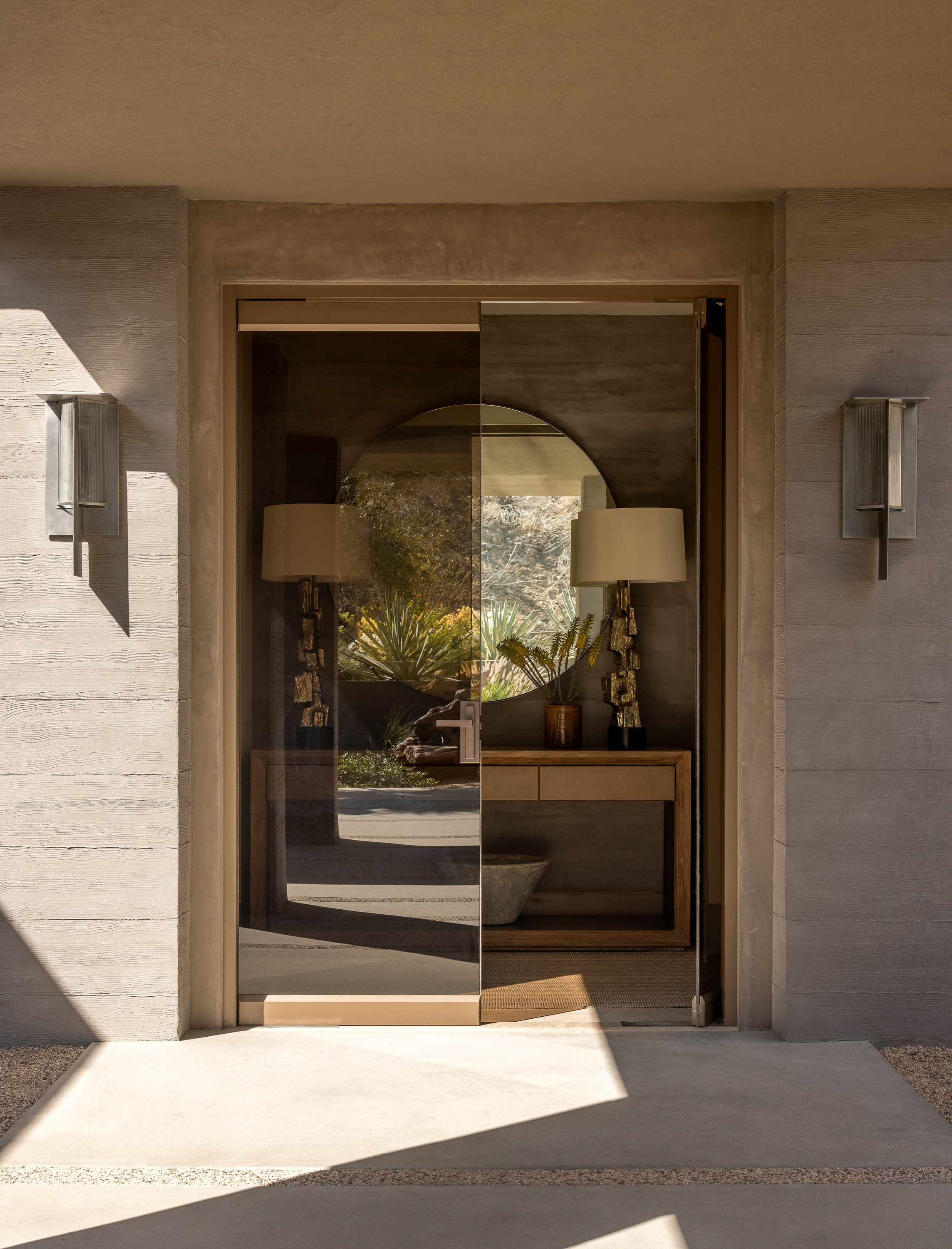 Grand entrance: concrete siding, double glass doors. One ajar, revealing brutalist lamps on an entry console and a giant circular mirror above.