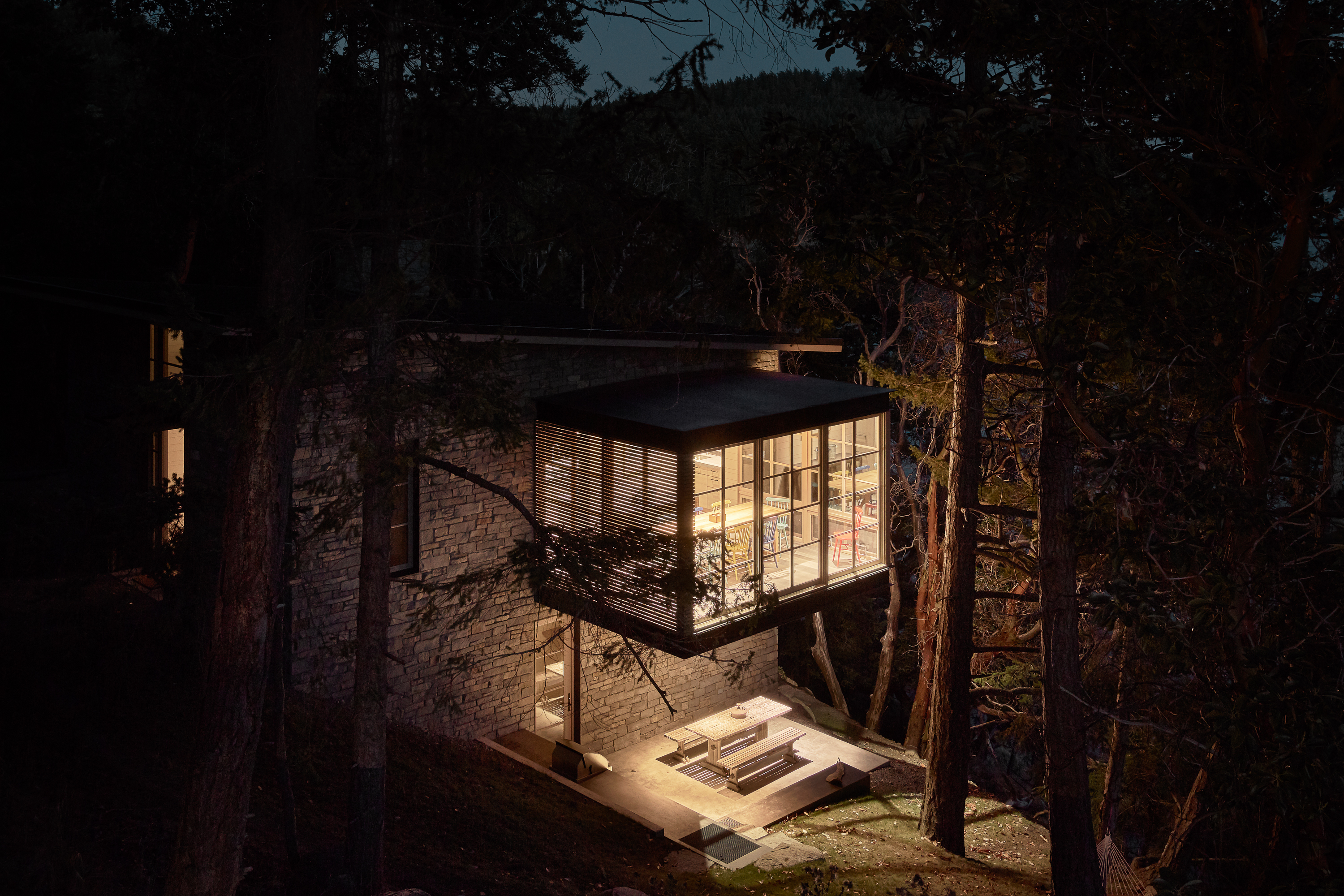 The house glows warmly from within, highlighting the cantilevered dining room that seemingly floats among the trees, creating a magical evening ambiance.